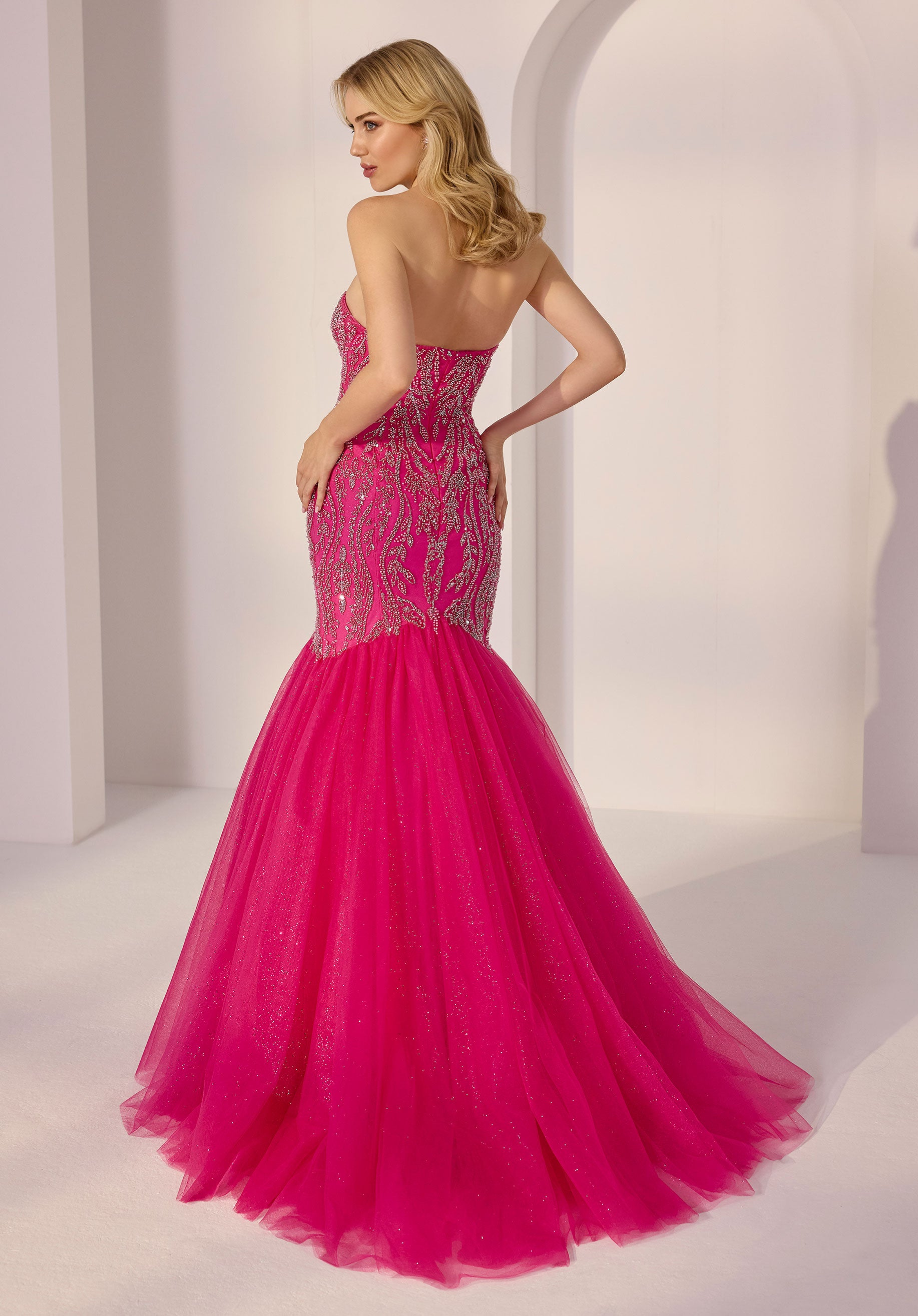 A woman wears the Strapless Mermaid Prom Dress: Morilee 49203 with a sequin bodice, standing facing away in a softly lit, minimalist setting.
